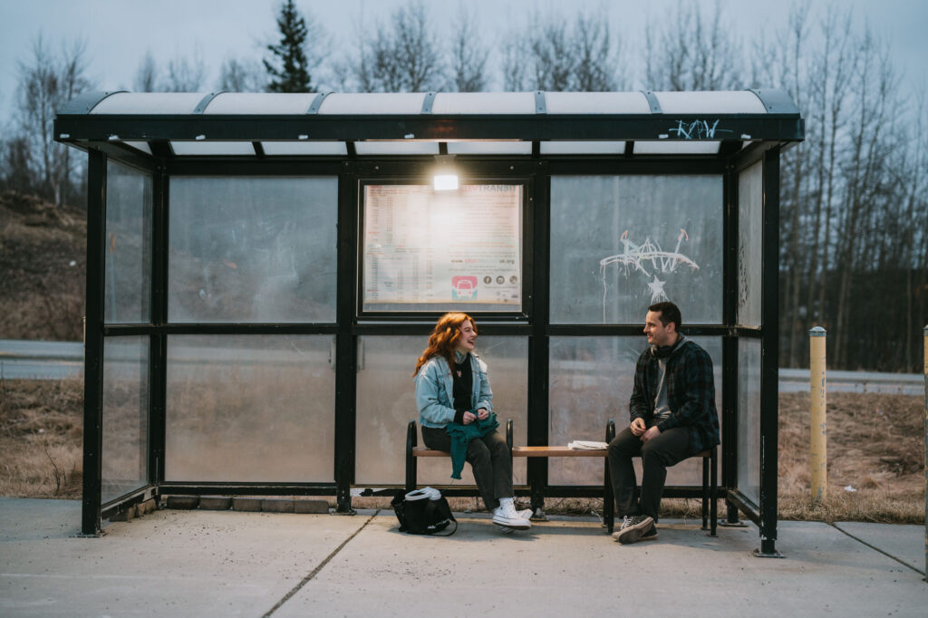 Two people sit on a bench inside a glass bus shelter, facing each other and talking. There is a backpack on the ground and trees in the background.