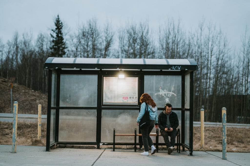 Two people wait at a bus stop shelter; one stands while the other sits looking at a phone. The shelter is surrounded by bare trees and has graffiti on the glass.