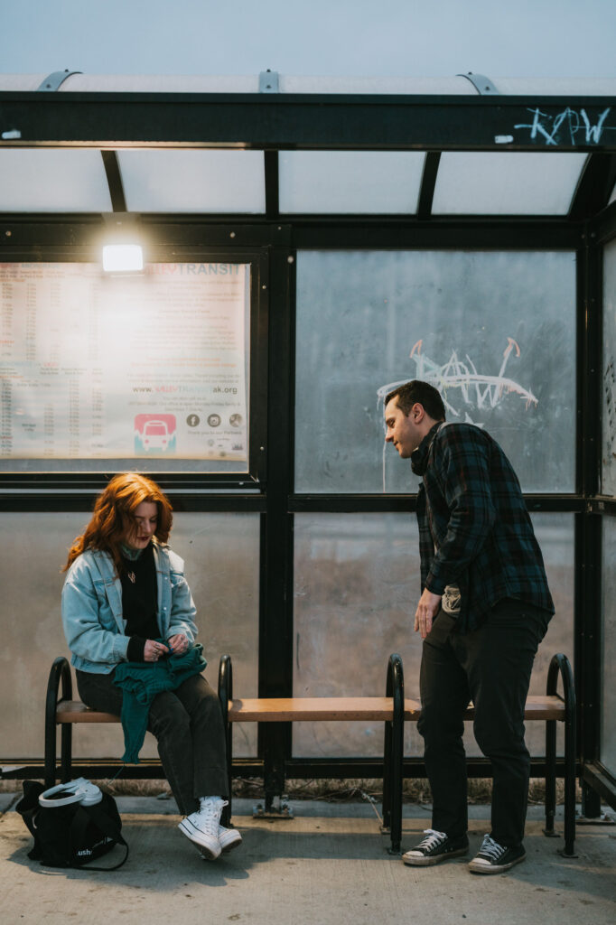 A woman sits on a bench at a bus stop, looking down, while a man stands nearby leaning towards her. It is evening and the bus stop is partially covered with glass panels.