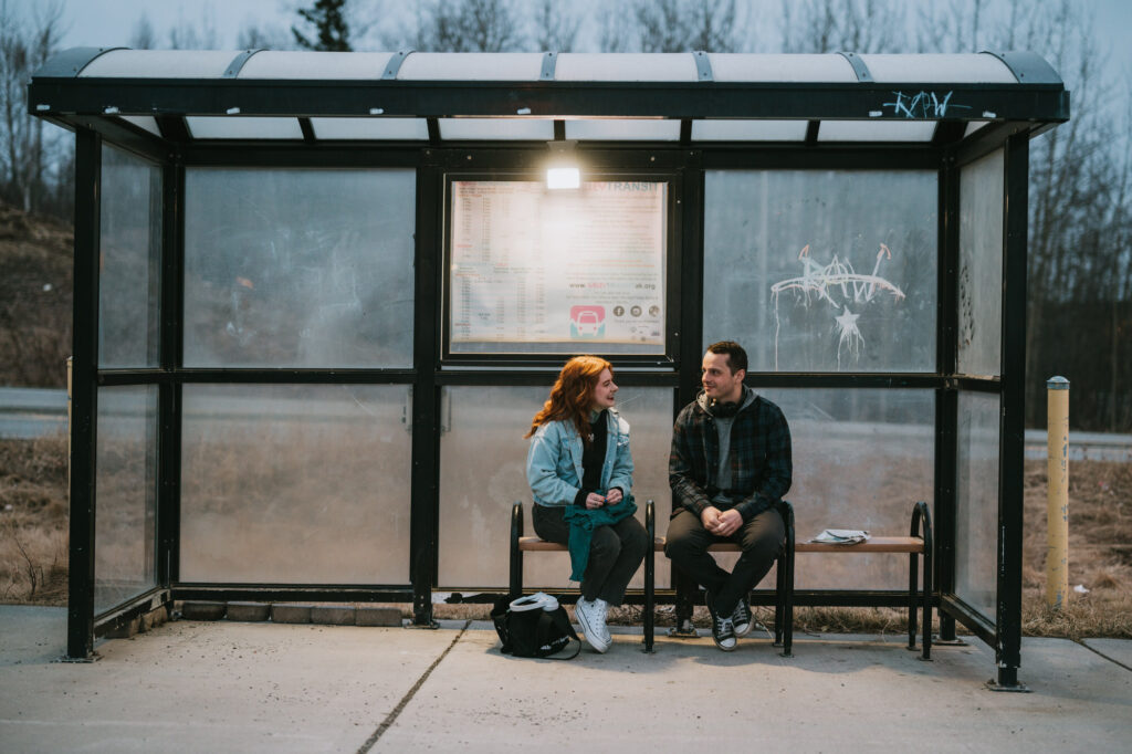 Two people sit and talk on a bench inside a mostly empty bus shelter, with a backpack on the ground and transit schedule posted behind them. It appears to be late afternoon or early evening.