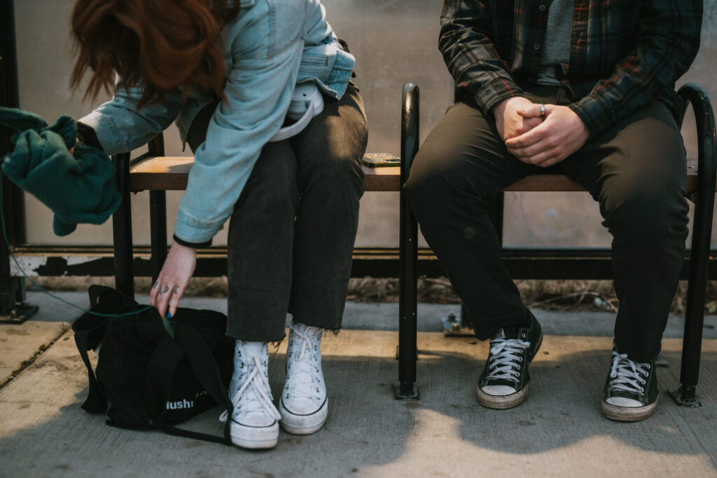 Two people sit on a bench; one is bending over to reach into a black bag, while the other sits with hands clasped, both wearing casual clothes and sneakers.