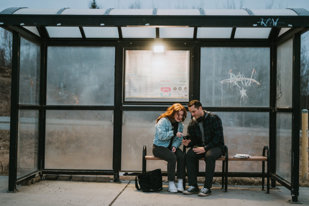 Two people sit closely together on a bench at a bus stop, looking at a smartphone and smiling. It is evening, and a backpack rests on the ground nearby.