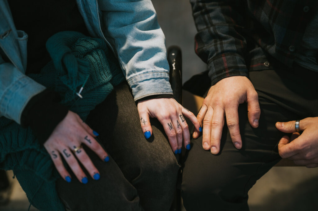 Two people sit side by side, one with blue nail polish and finger tattoos reading "SELF MADE," their hands resting on their laps and touching.