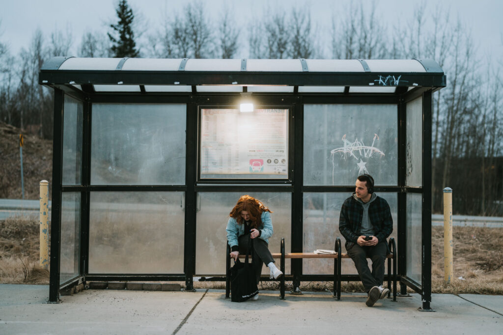 Two people sit apart on a bench at a bus stop; one looks at a phone, and the other looks down, tying a shoe. The setting is outdoors with leafless trees in the background.