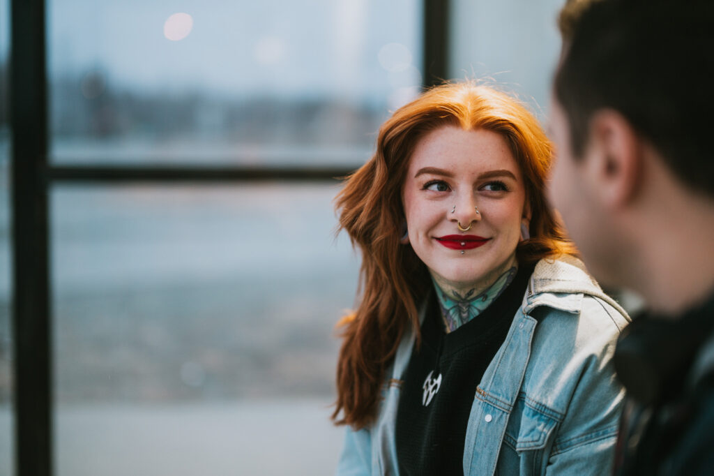 A woman with red hair, tattoos, and facial piercings smiles at a person sitting next to her indoors by a window.