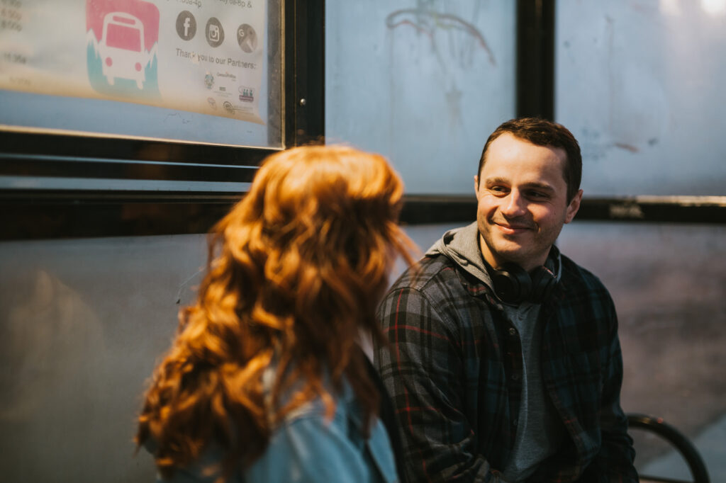 A man with headphones around his neck smiles at a woman with red hair while sitting indoors near a window with posters.