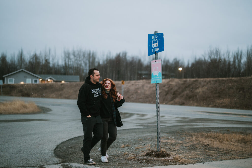 A man and woman walk together on a sidewalk by a bus stop sign on a cloudy day, with a house and trees in the background.