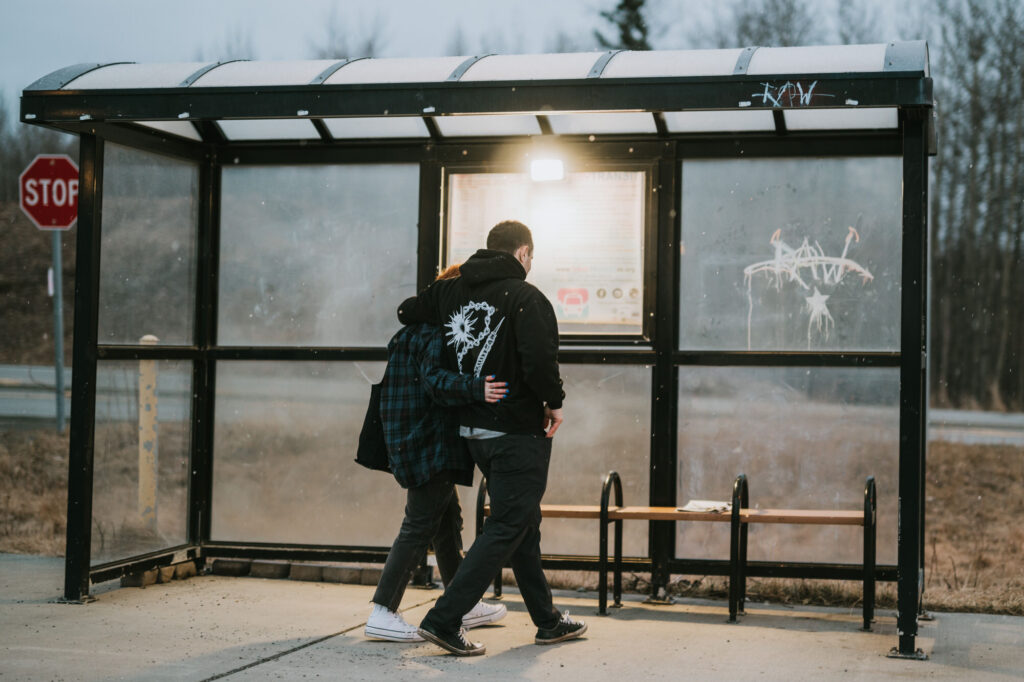 Two people stand side by side under a bus shelter, looking at a posted schedule; one has an arm around the other. The shelter is empty except for a bench and some graffiti.