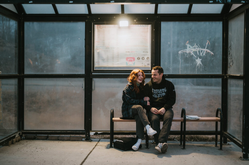 Two people sit closely on a bench at a bus stop, laughing together. It's evening, and the shelter is lit by an overhead light with a schedule posted behind them.