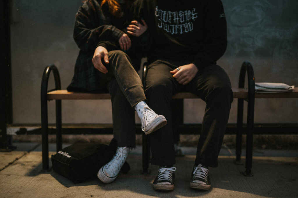 Two people sit closely together on a bench, one with an arm around the other. They wear casual clothing and sneakers. A backpack rests on the ground near their feet.