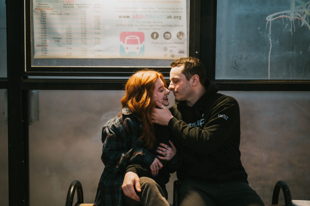 A man gently holds a woman's face as they sit close together on a bench, looking into each other's eyes with a schedule posted on the wall behind them.