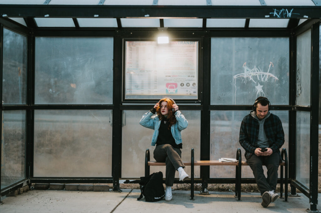 Two people sit on a bench at a bus stop. One person adjusts headphones, and the other looks at a phone. Both are dressed casually and the shelter has a schedule posted behind them.