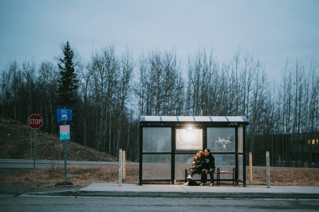 Two people sit together on a bench inside a glass bus shelter at dusk, surrounded by leafless trees and a stop sign nearby.