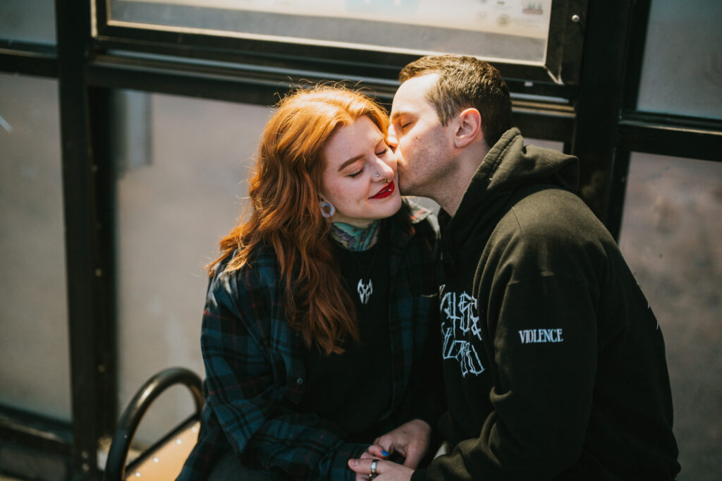 A man kisses a woman on the cheek while they sit close together on a bench. The woman smiles and looks ahead.