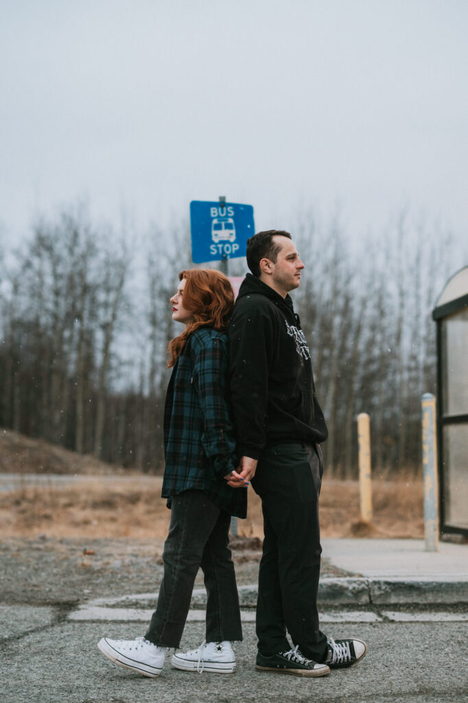 Two people stand back to back holding hands near a bus stop sign on a cloudy day, with trees and dry grass in the background.