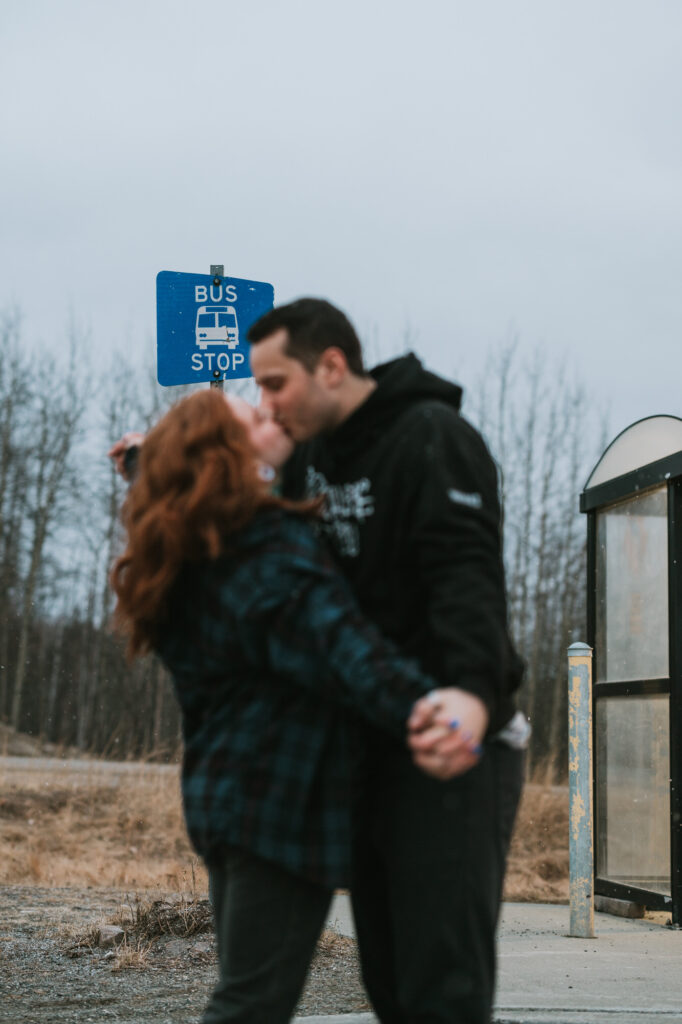 Two people kiss while standing in front of a bus stop sign and shelter on an overcast day, with barren trees and dry grass in the background.