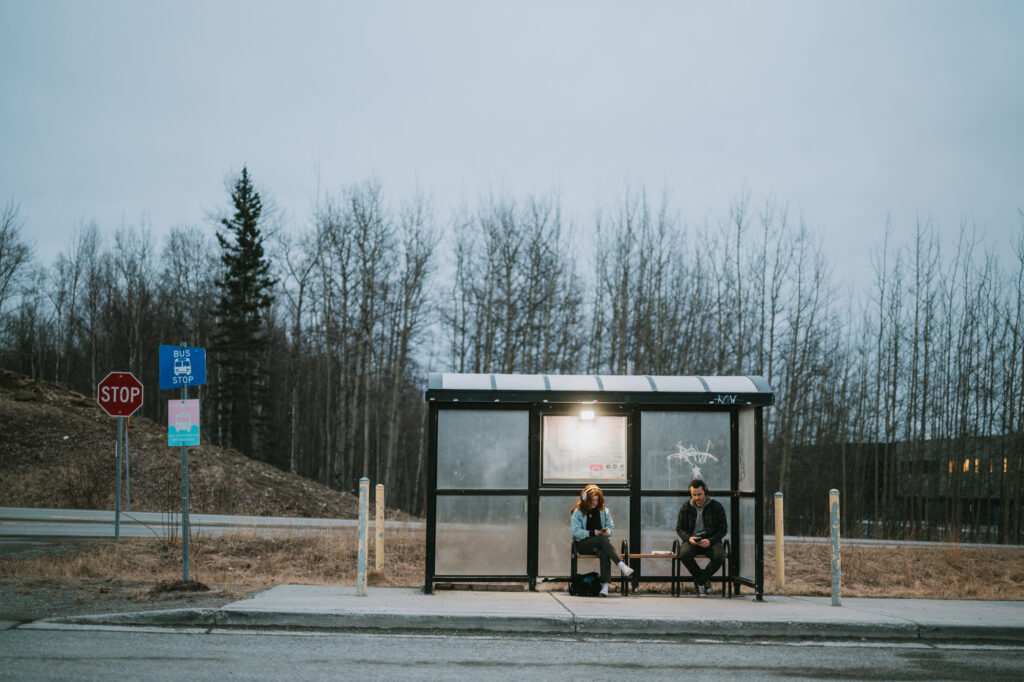 Two people sit inside a glass bus shelter at dusk, with bare trees and a stop sign in the background on a quiet roadside.