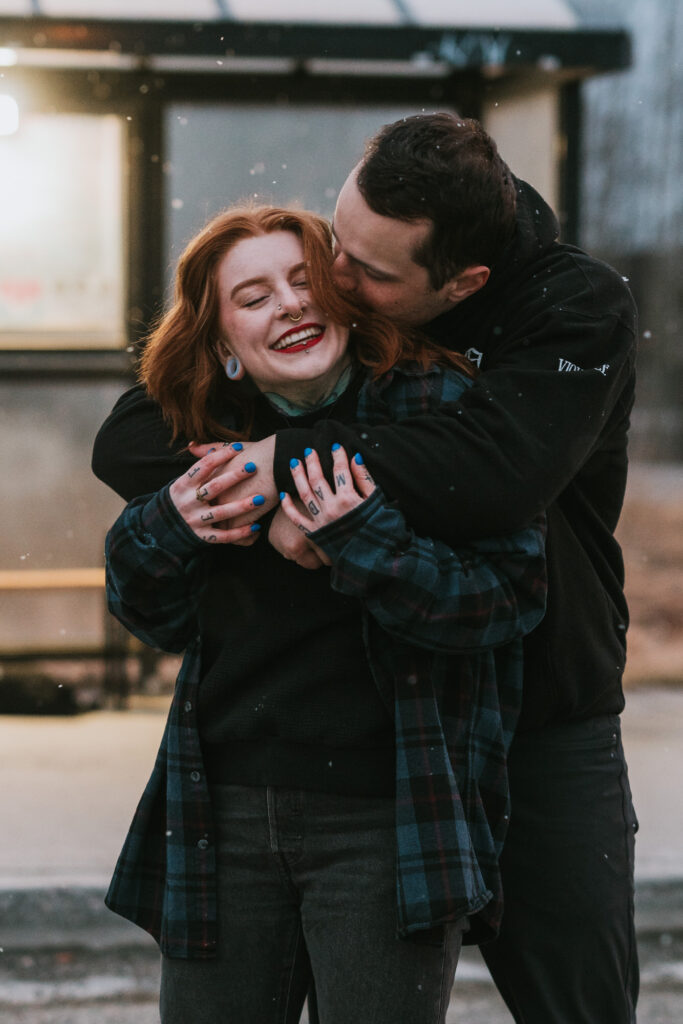 A man embraces a smiling woman from behind outdoors, both dressed in casual clothing, with snowflakes visible in the air.