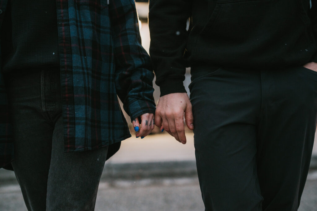 Two people wearing dark clothing are holding hands while walking outdoors. Only their torsos, arms, and hands are visible.