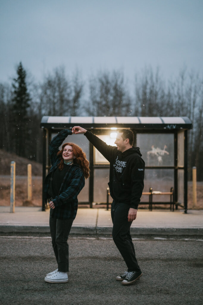Two people dance together in front of a bus shelter on a cloudy day, with trees and a light in the background.