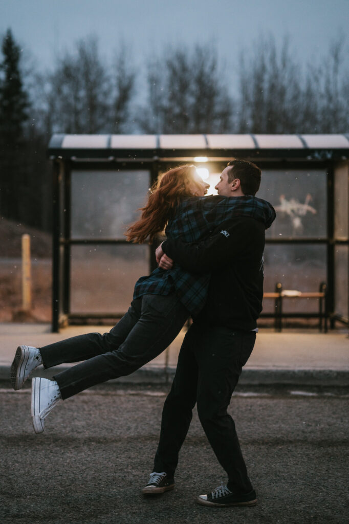 A person lifts and spins another person in front of a bus stop at dusk, both smiling and dressed in casual clothing. Trees and a lit bus shelter are in the background.