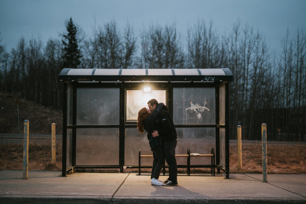 A couple stands and kisses under a lit bus shelter at dusk; leafless trees and a road are visible in the background.