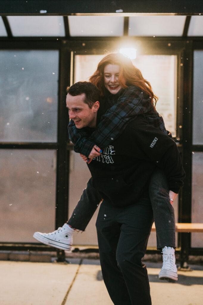 A man gives a woman a piggyback ride outside under a covered shelter; both are smiling and dressed in casual clothing.