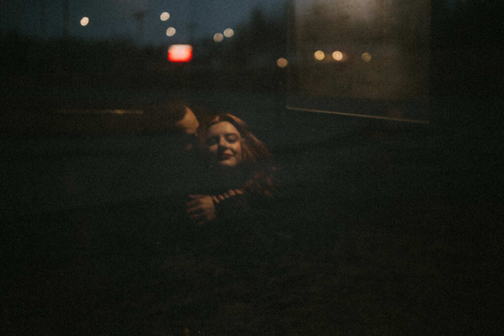 A couple hugs closely at night, illuminated by soft streetlights and distant blurry lights, creating a dark, intimate atmosphere.