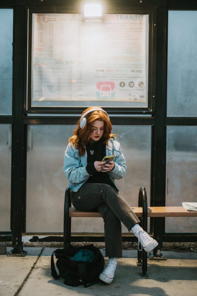 A person with headphones sits on a bench at a transit stop, looking at their phone. A schedule is posted behind them, and a backpack rests on the ground nearby.