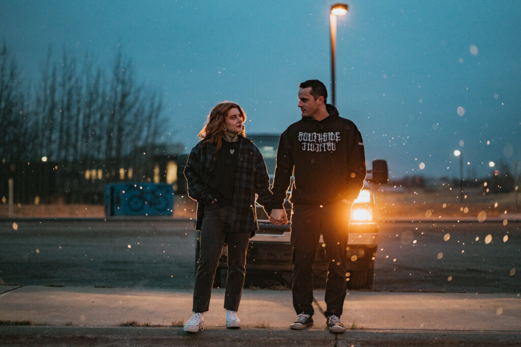 Two people stand hand in hand on a sidewalk at dusk, illuminated by a car’s headlights with falling snow and bare trees in the background.