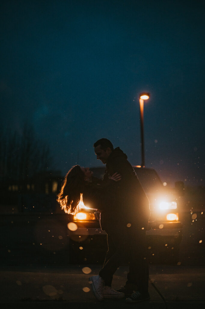 A couple stands close together at night in front of a car with headlights on, with one person leaning back and the other supporting them. A streetlight is visible in the background.