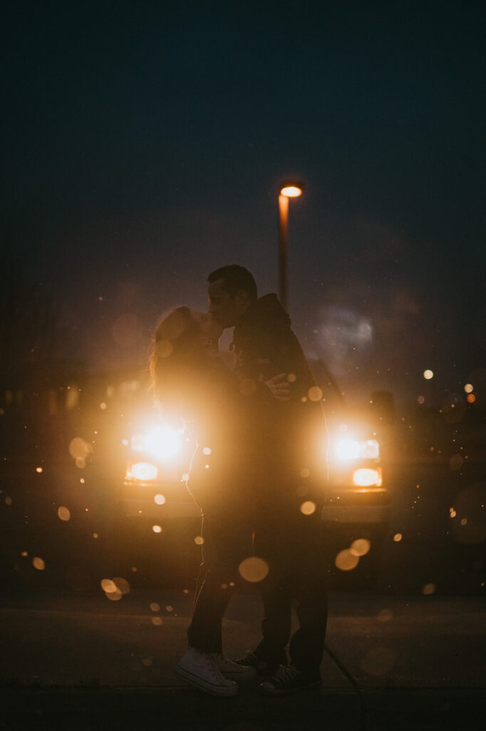 A couple kisses in front of a vehicle with headlights on at night, silhouetted by the lights with raindrops visible in the foreground.
