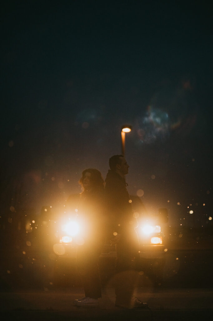 Two people stand back-to-back in front of a car with headlights on at night, illuminated by the car lights, with bokeh effects visible in the foreground.