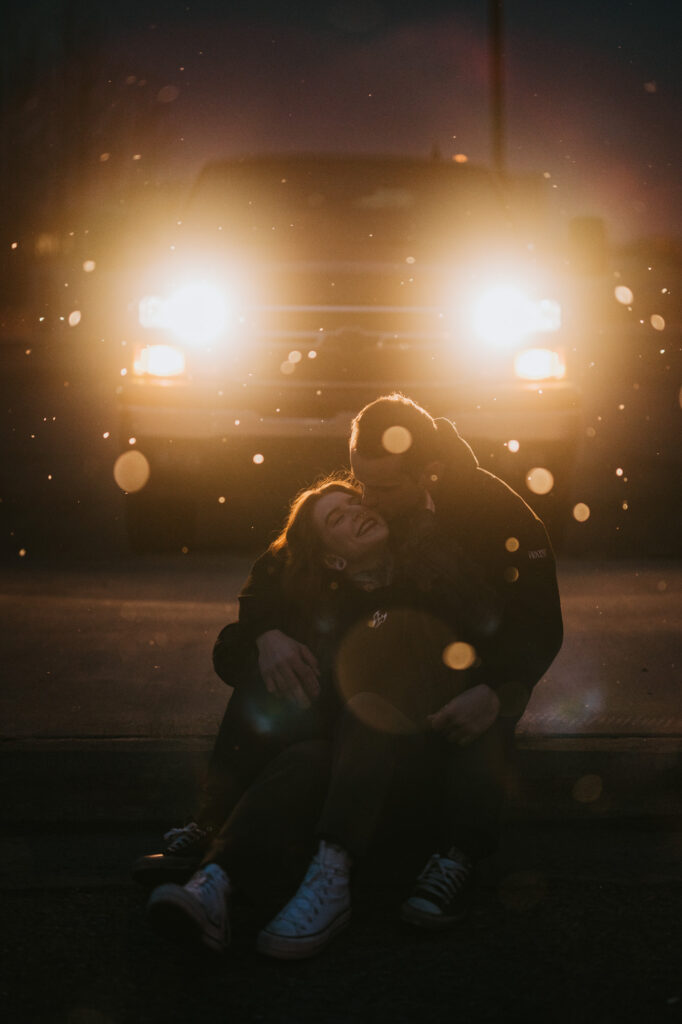 Two people sit close together on the ground at night in front of a vehicle with its headlights on, with light flares and particles visible in the air.