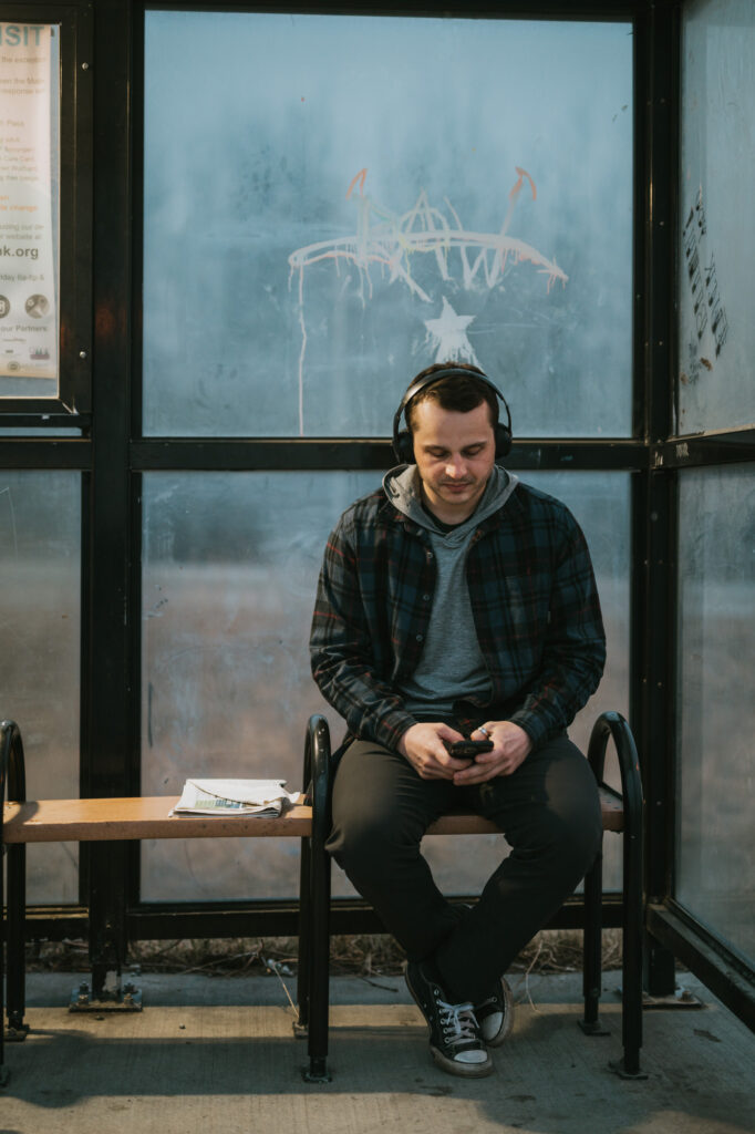 A man wearing headphones sits on a bench at a bus stop, looking at his phone. A folded newspaper is on the bench beside him. The glass behind him is marked with graffiti.
