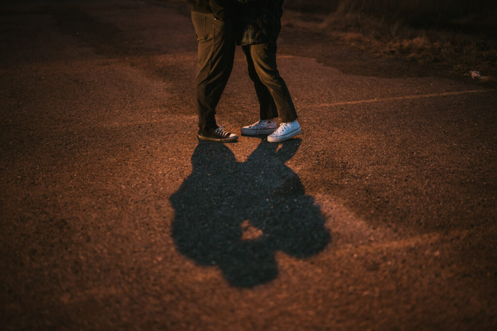 Two people standing close together on a paved surface, their legs and shoes visible, casting a shadow in the warm outdoor light.