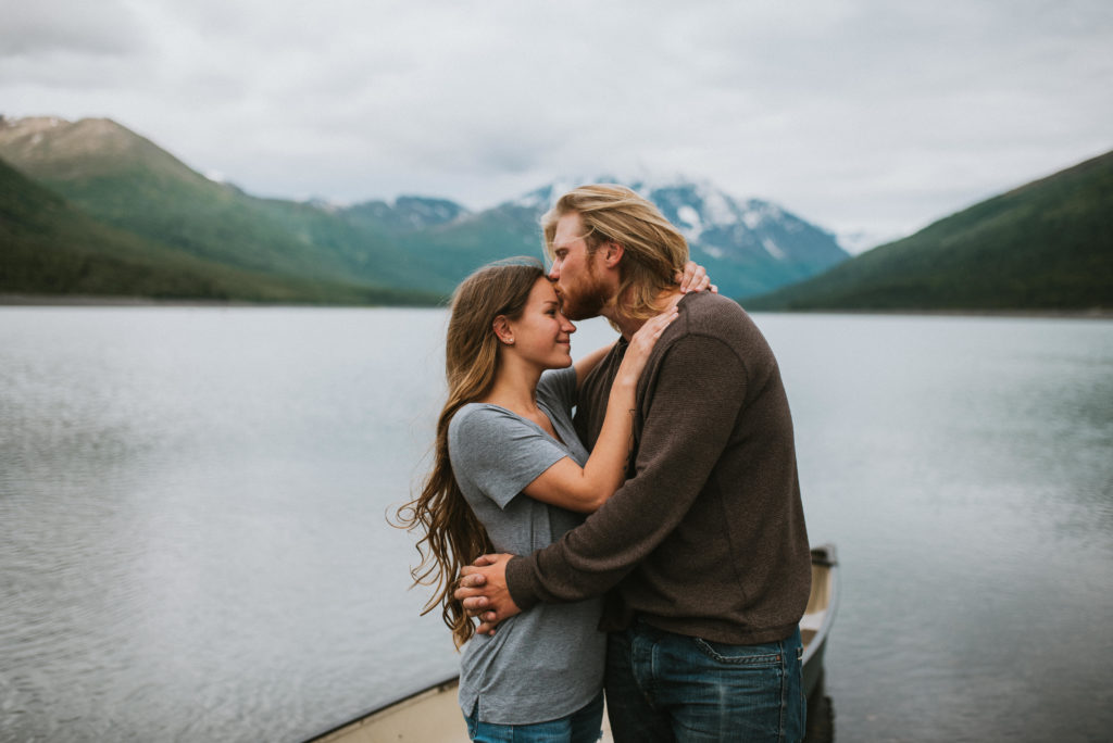 Couple hugging on lake shore with mountain backdrop, he is kissing her forehead